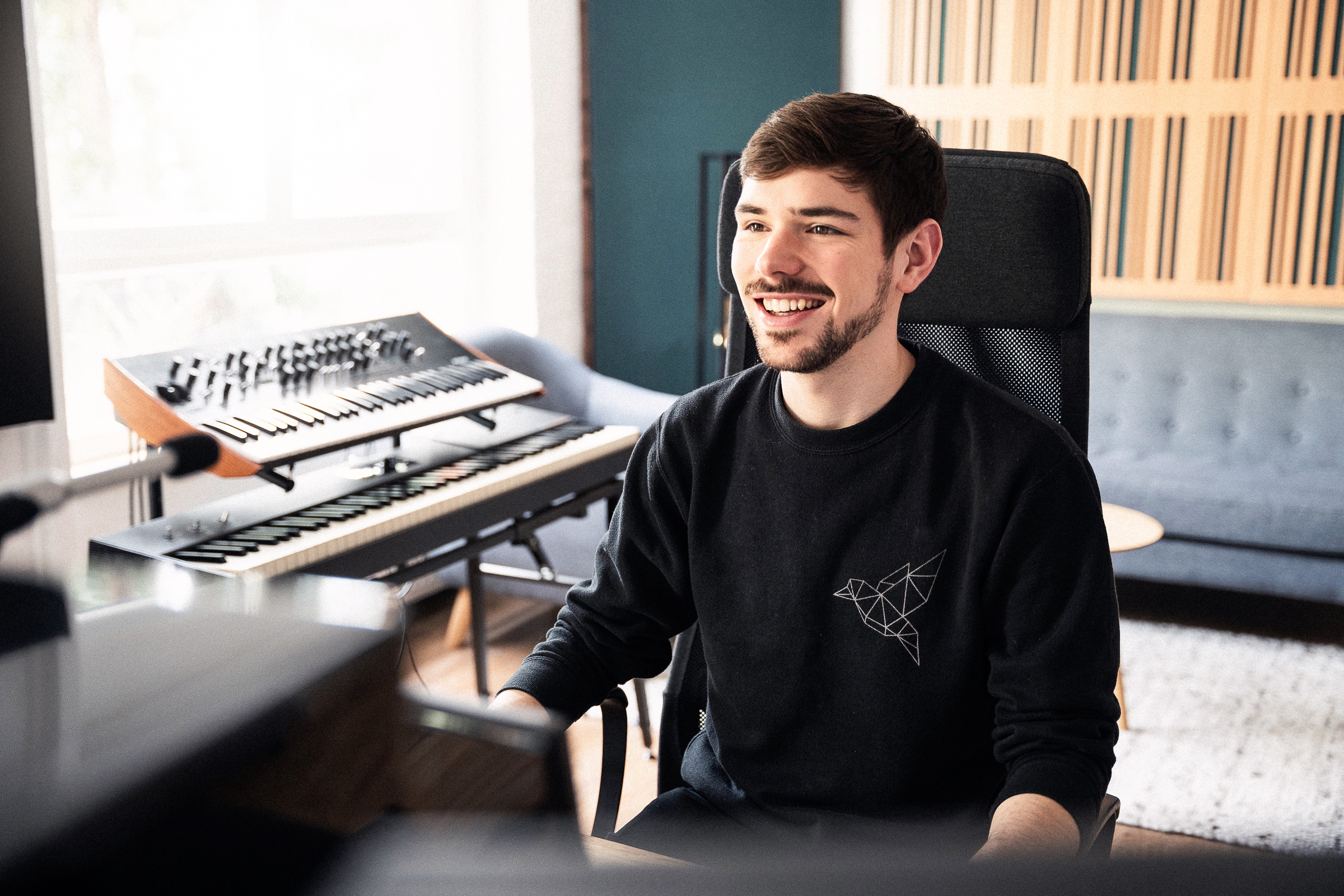 Sam sitting in the control room in front of keyboards and a sofa